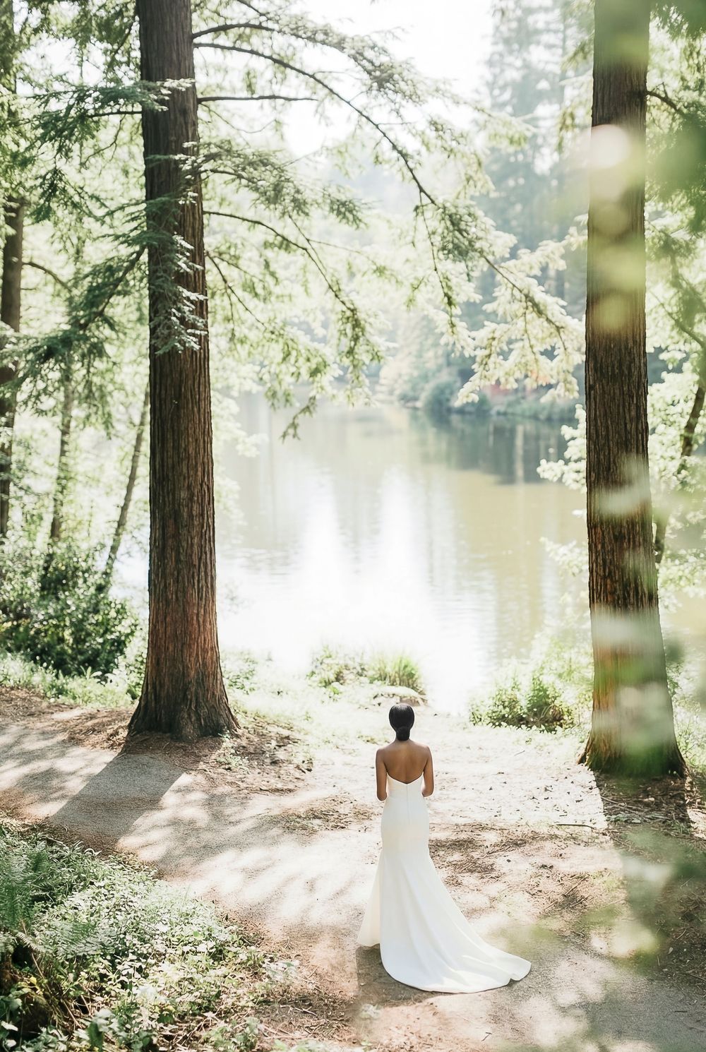 Bride in white gown standing between tall trees overlooking a serene forest lake on her wedding day.