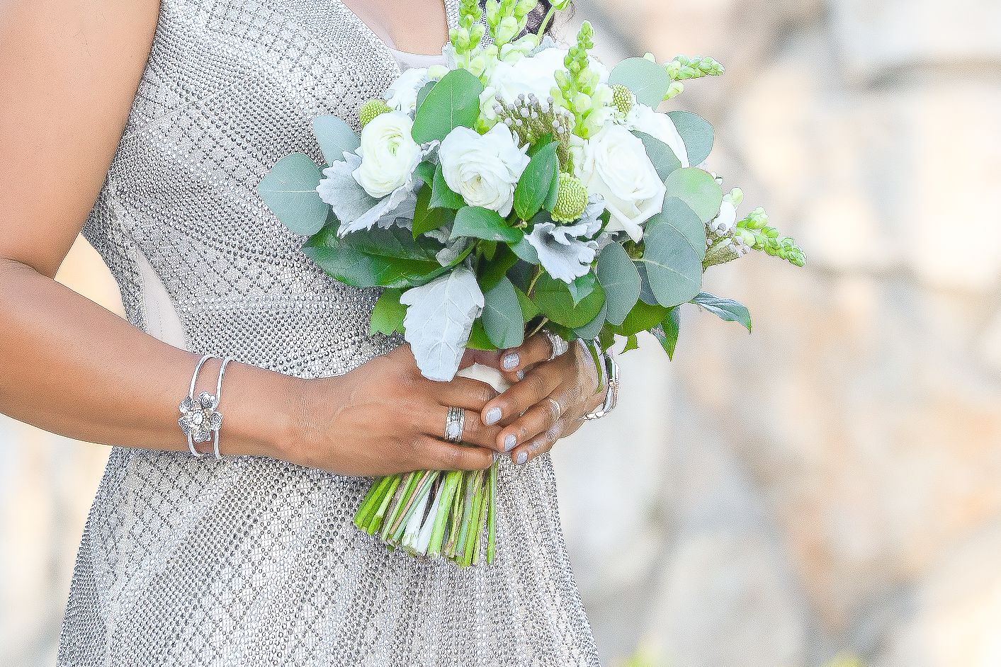 Bride holding white floral bouquet with greenery, wearing silver beaded dress and diamond bracelet.