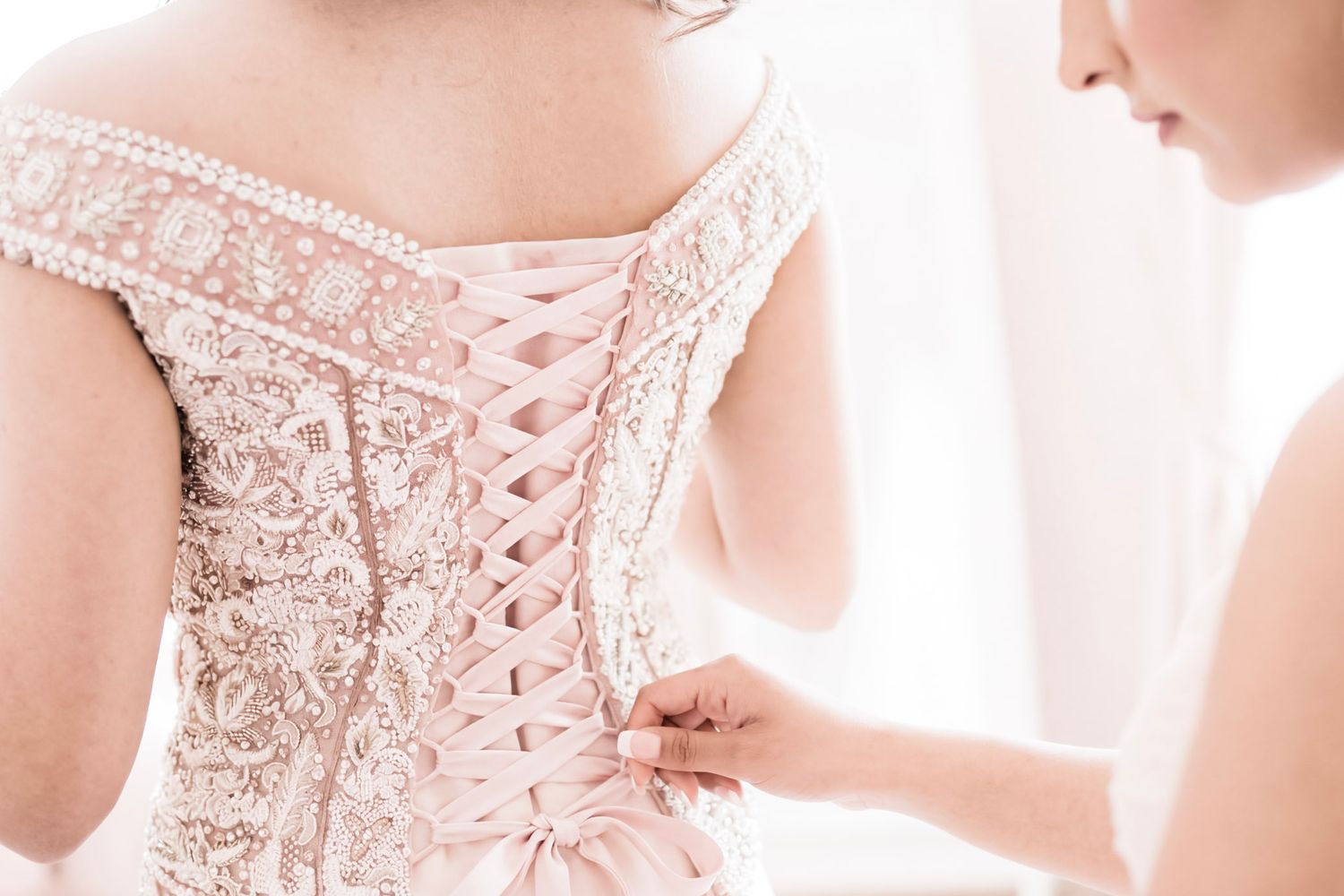 Bridesmaid lacing up the back of a blush pink beaded off-shoulder wedding dress.