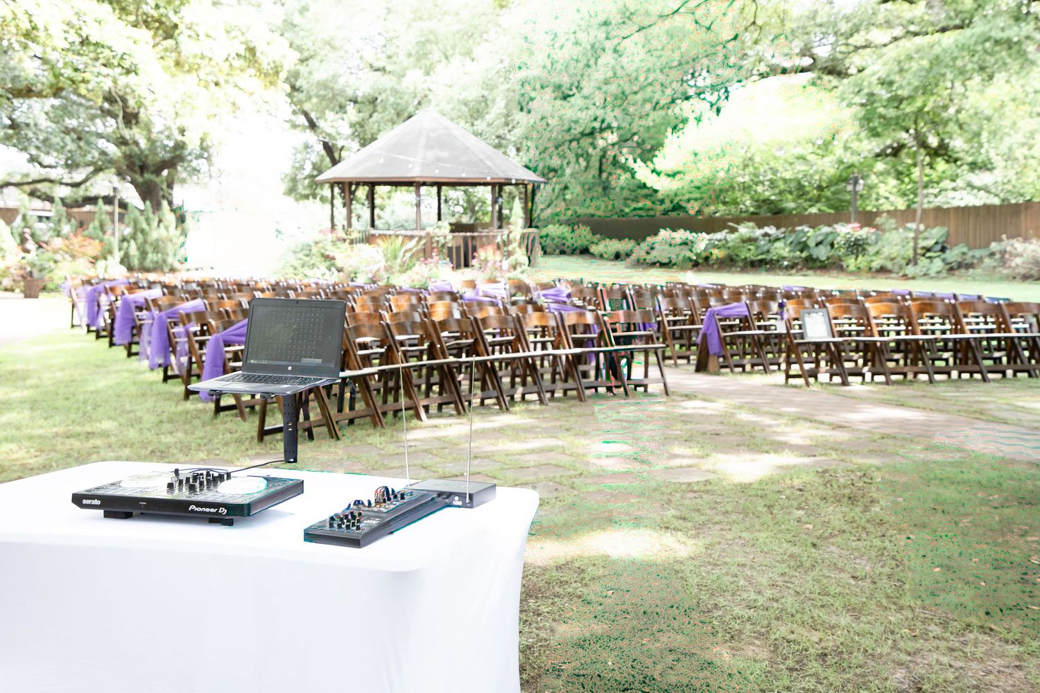 Outdoor wedding ceremony setup with wooden chairs, purple accents, DJ equipment in foreground, and a gazebo amid lush greenery.