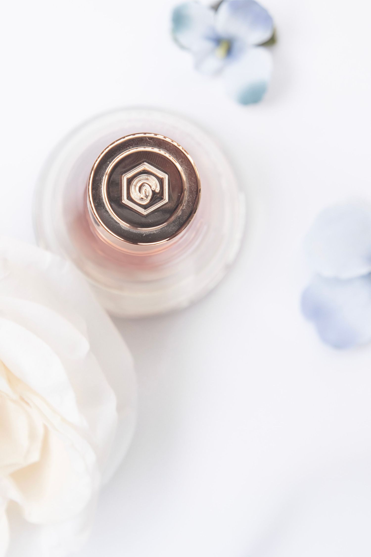 A rose gold perfume bottle cap surrounded by delicate white roses and soft blue flowers on a white background.