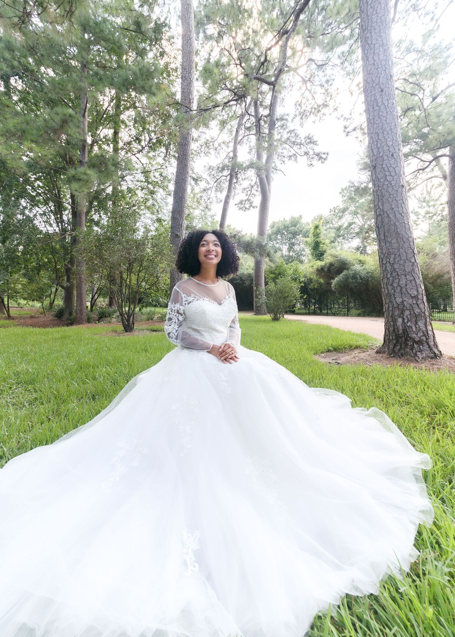 A bride in a flowing white ballgown twirls gracefully in a sunlit wooded area with tall pine trees and lush grass.