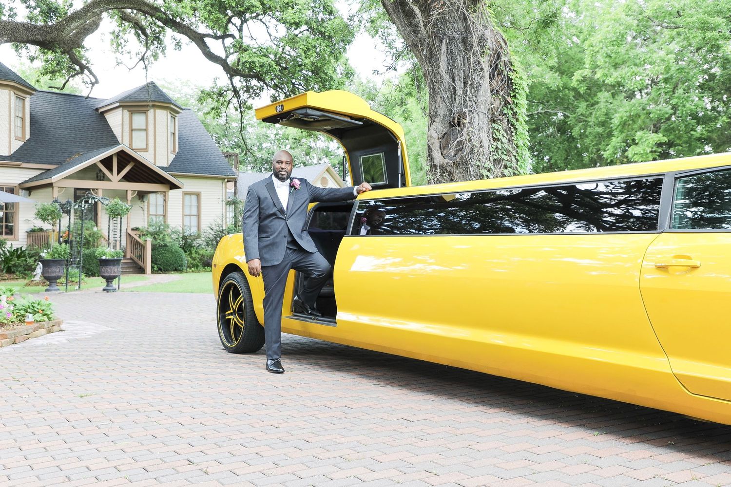 Man in suit stepping out of a bright yellow stretch limousine on a brick driveway near a historic home.