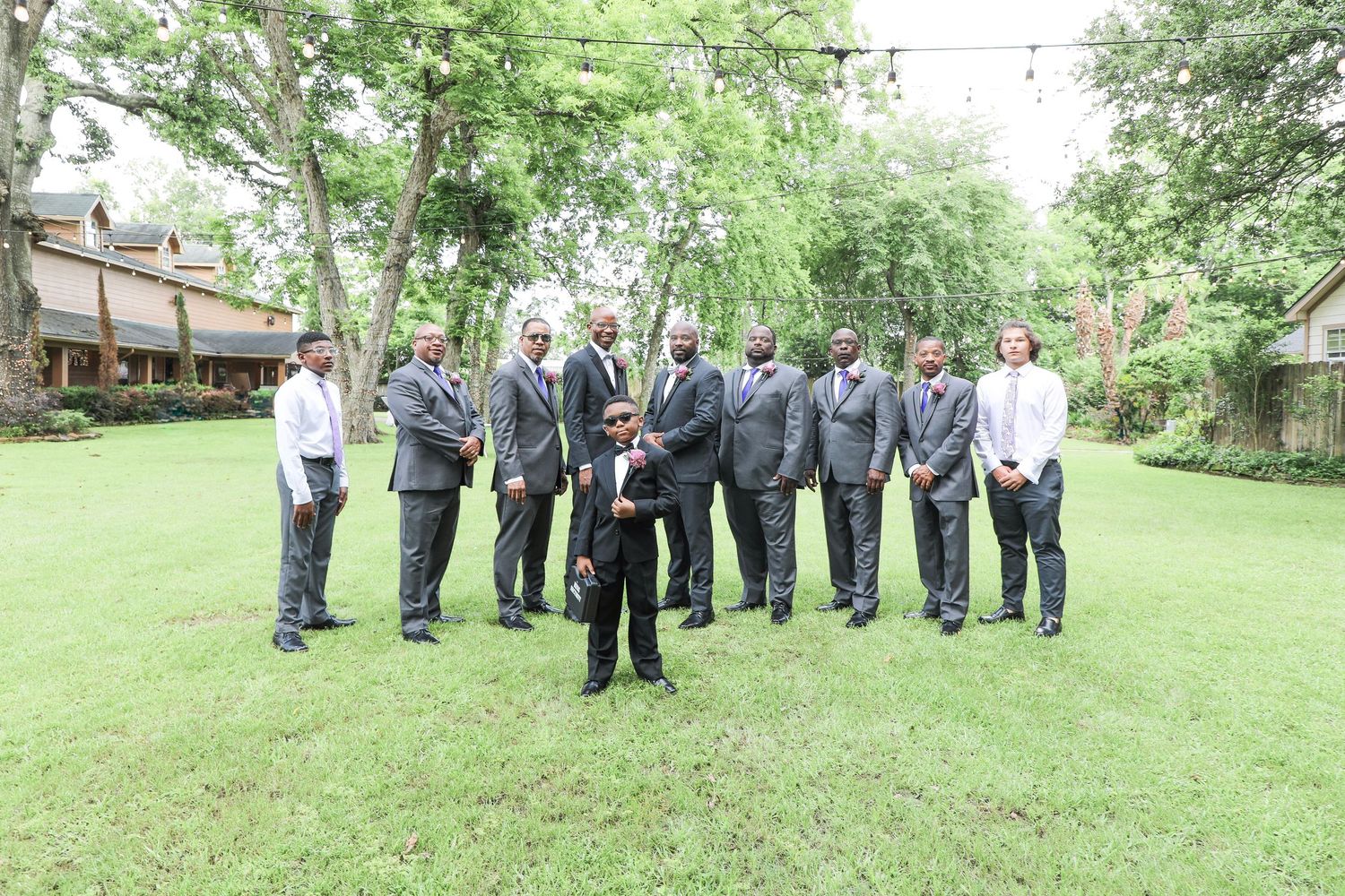 Group of men in gray suits posing outdoors on green lawn with trees and building in background.