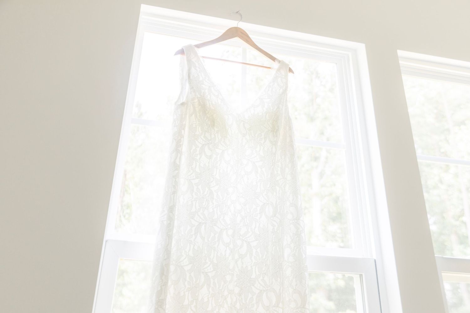 White lace wedding dress hanging on a wooden hanger in front of bright windows with natural light.