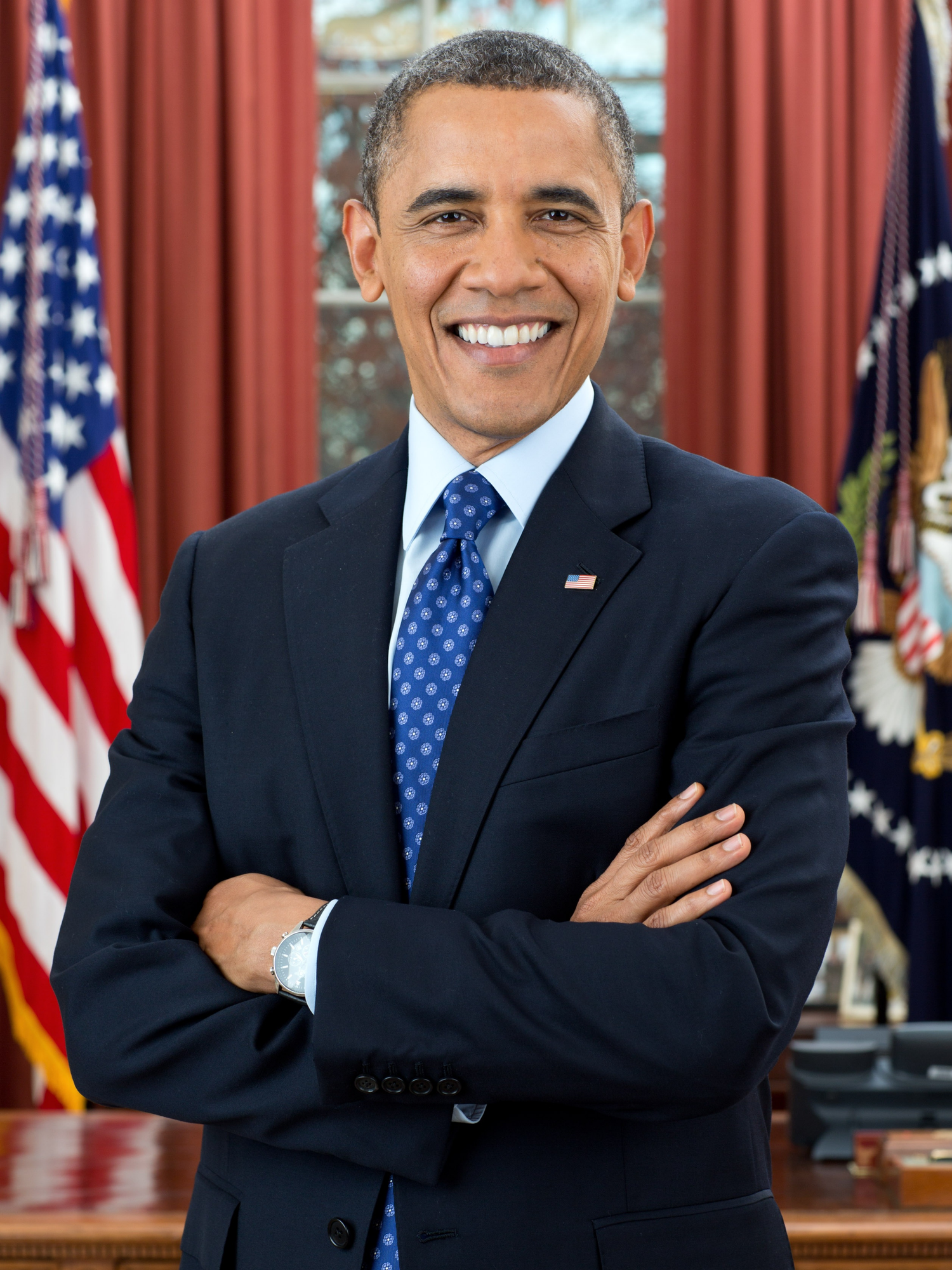 A figure in a dark suit and polka dot tie poses in front of American flags in the Oval Office.