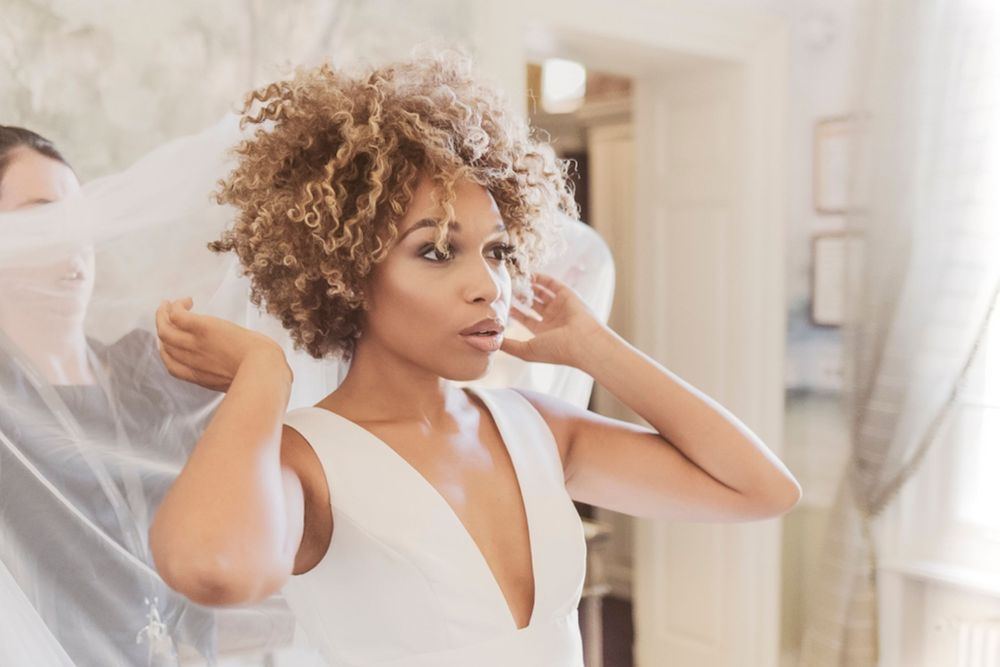 Person with curly hair wearing a white dress adjusts bridal veil in a softly lit room during wedding preparations.