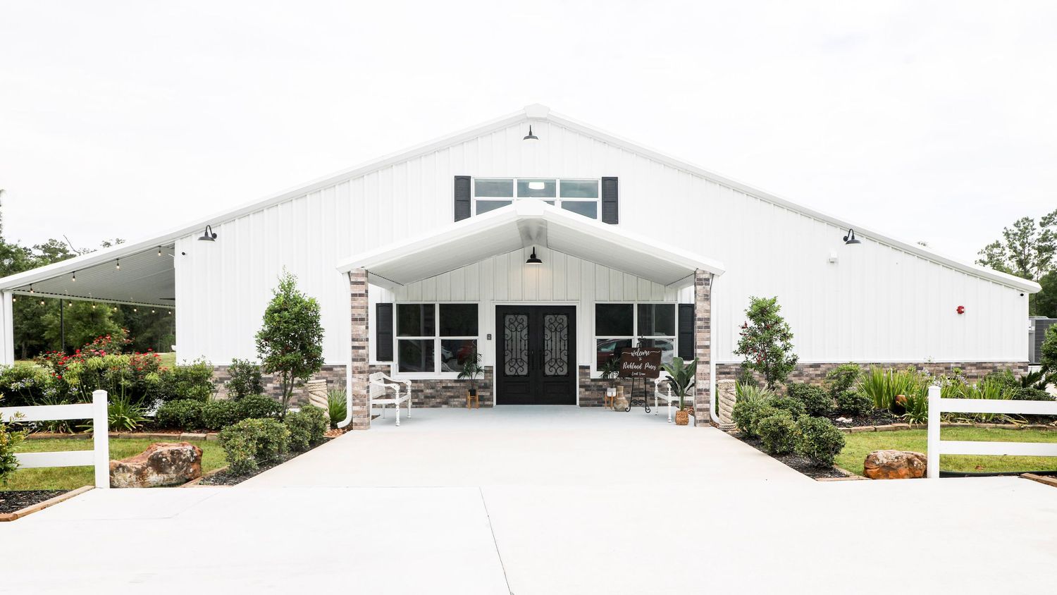 White barn-style wedding venue with stone accents, black shutters, and a paved entrance surrounded by lush greenery.