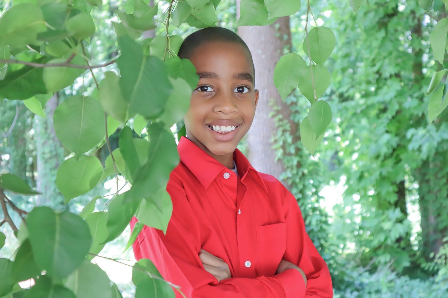 A child wearing a bright red jacket smiles while standing among green leafy foliage outdoors.