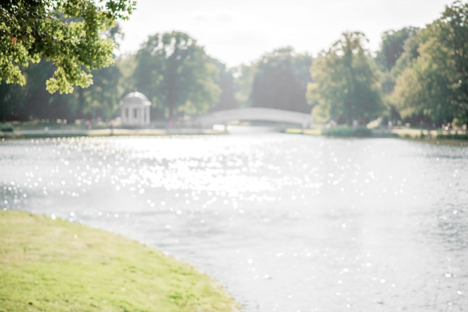 Sunlit lake with gazebo in the distance surrounded by trees and grassy shoreline on a misty summer morning.