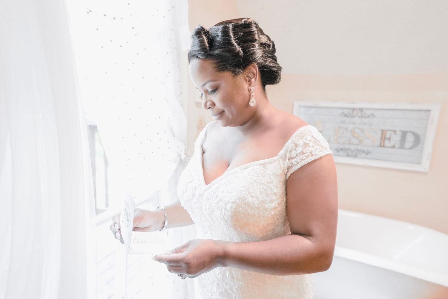 A bride in a white lace wedding dress stands near a window, reading a letter in an intimate moment on her wedding day.