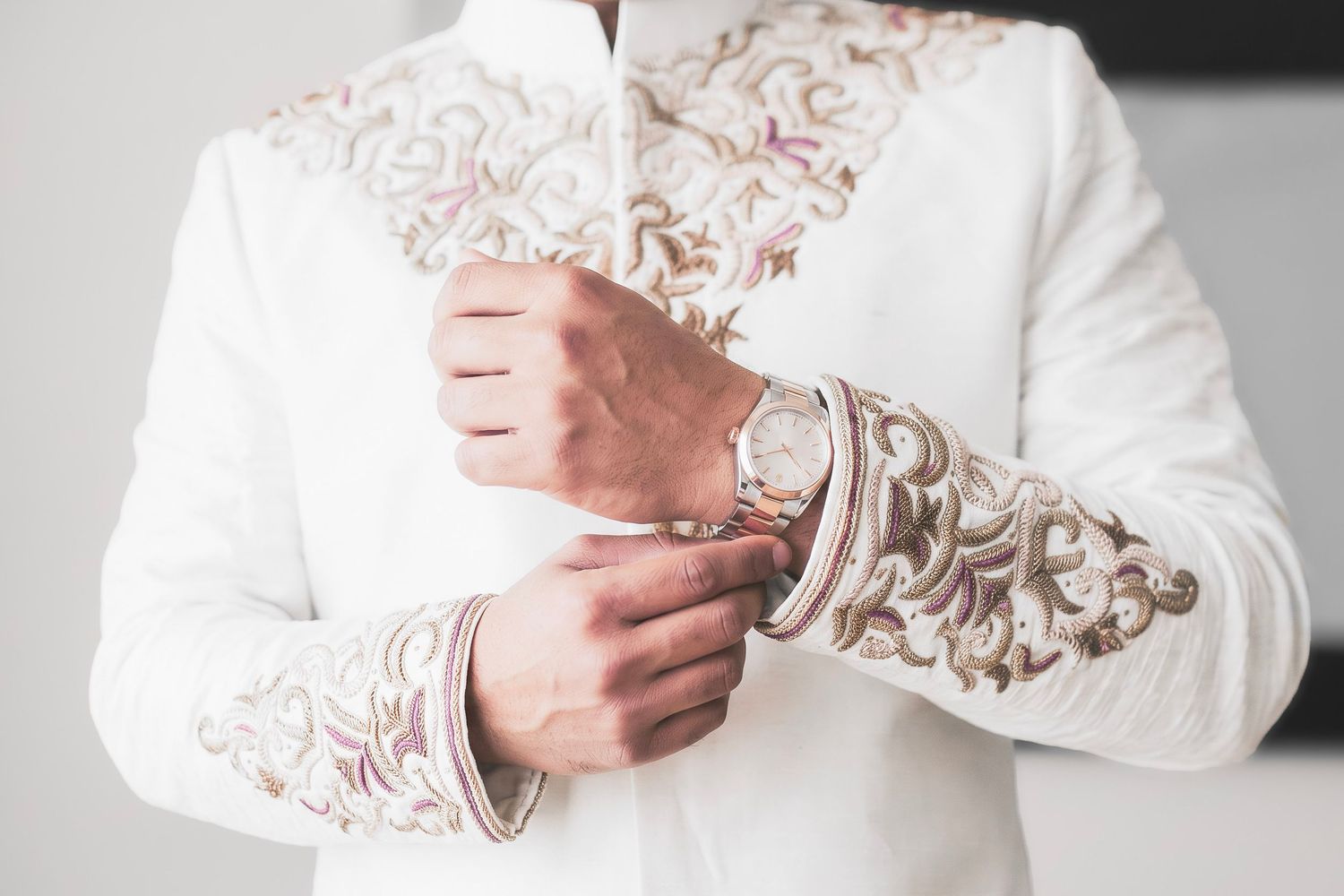 Man in white embroidered sherwani adjusting cufflinks, wearing a gold watch, elegant Indian wedding attire.