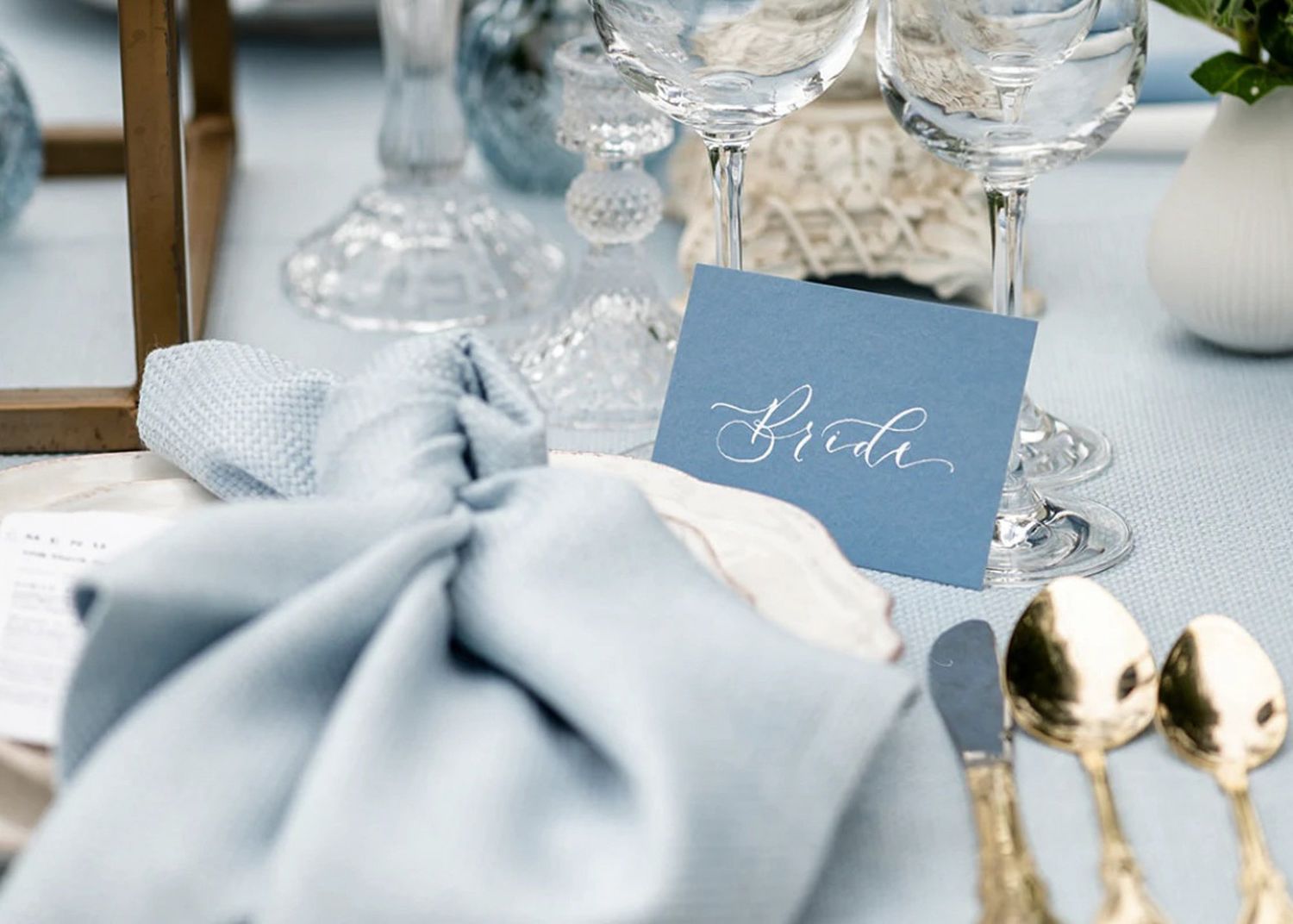 Elegant wedding place setting with dusty blue napkin, calligraphy name card, crystal glassware and gold cutlery.