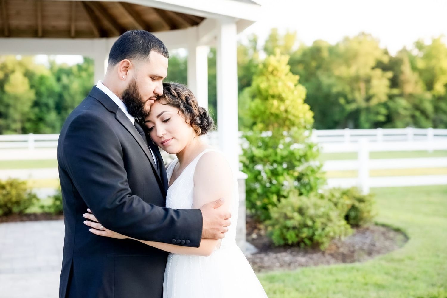 A bride and groom share a tender embrace outdoors near a white fence and gazebo on their wedding day.