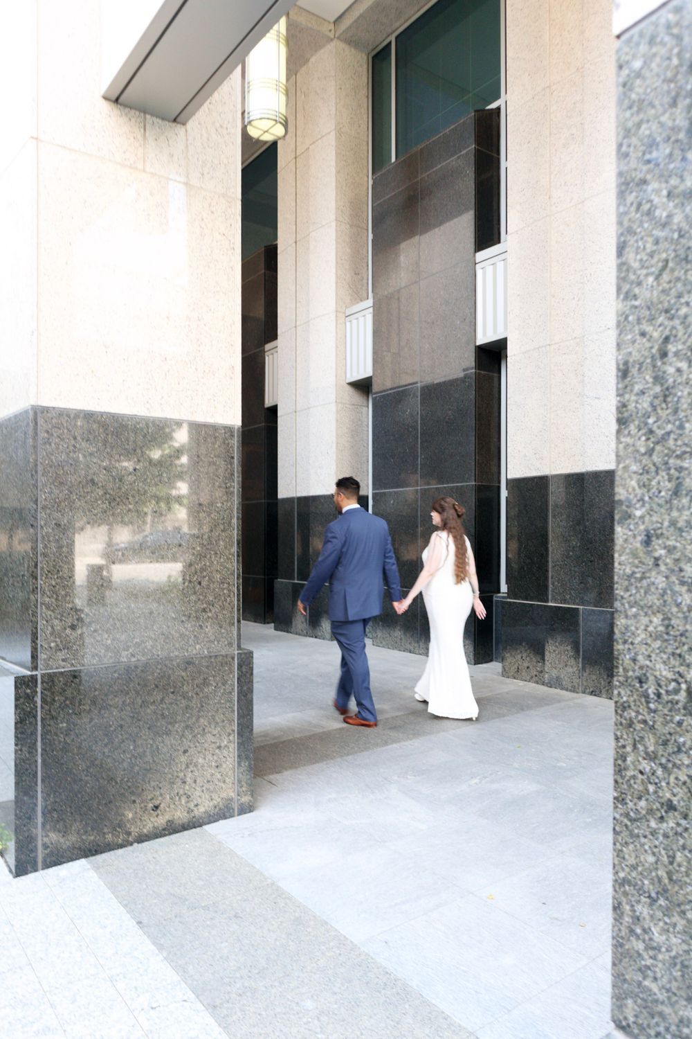 Bride and groom walking hand in hand toward a modern building entrance on their wedding day.