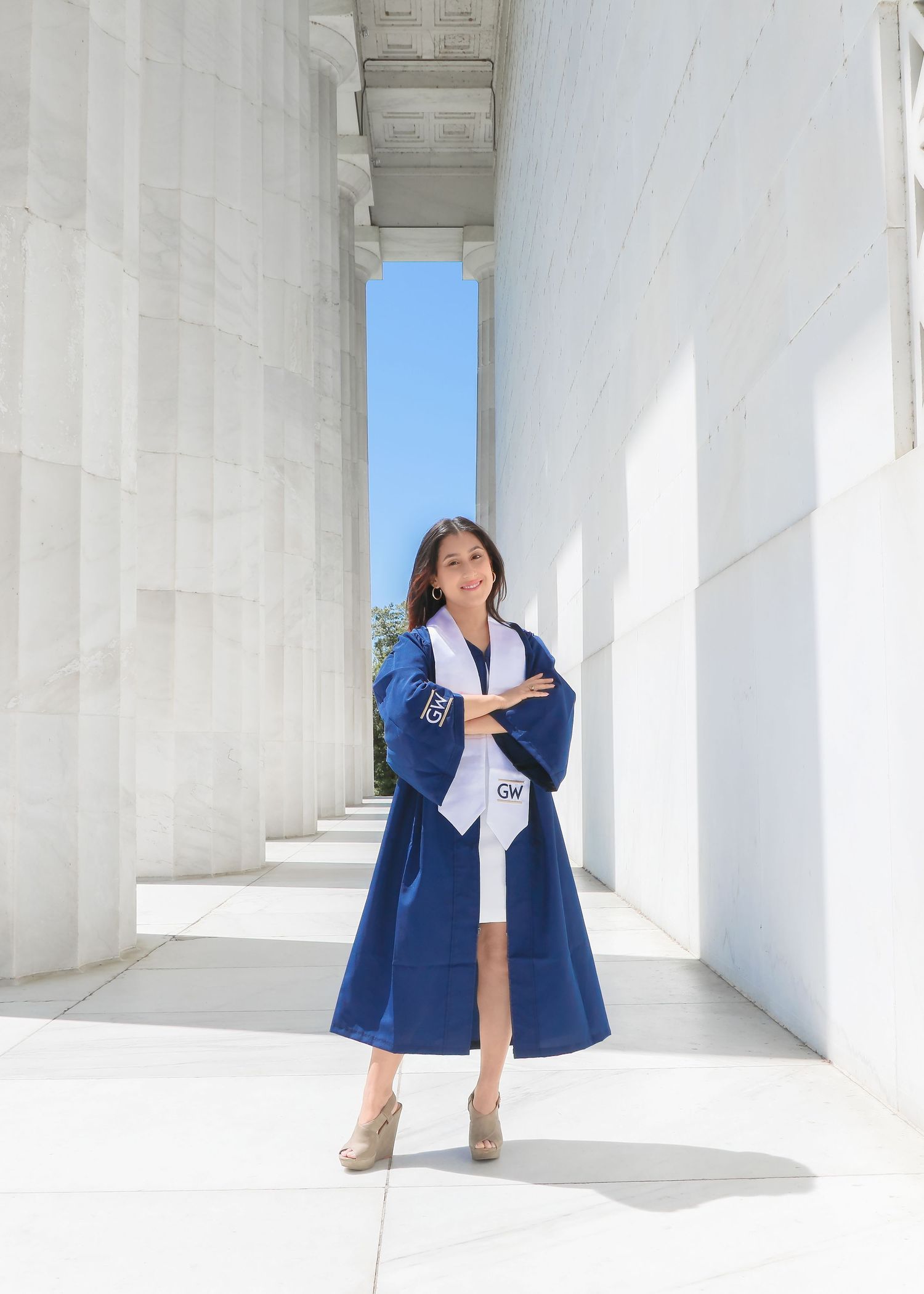 Someone in blue graduation regalia stands posing between white marble columns with blue sky visible.