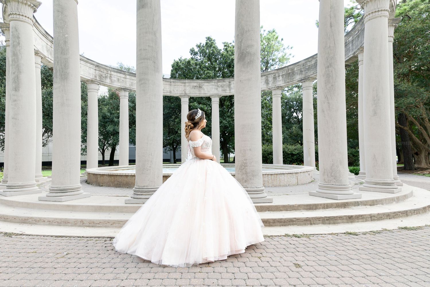 A panoramic view of white marble columns in a classical garden setting with a figure in a flowing white dress.