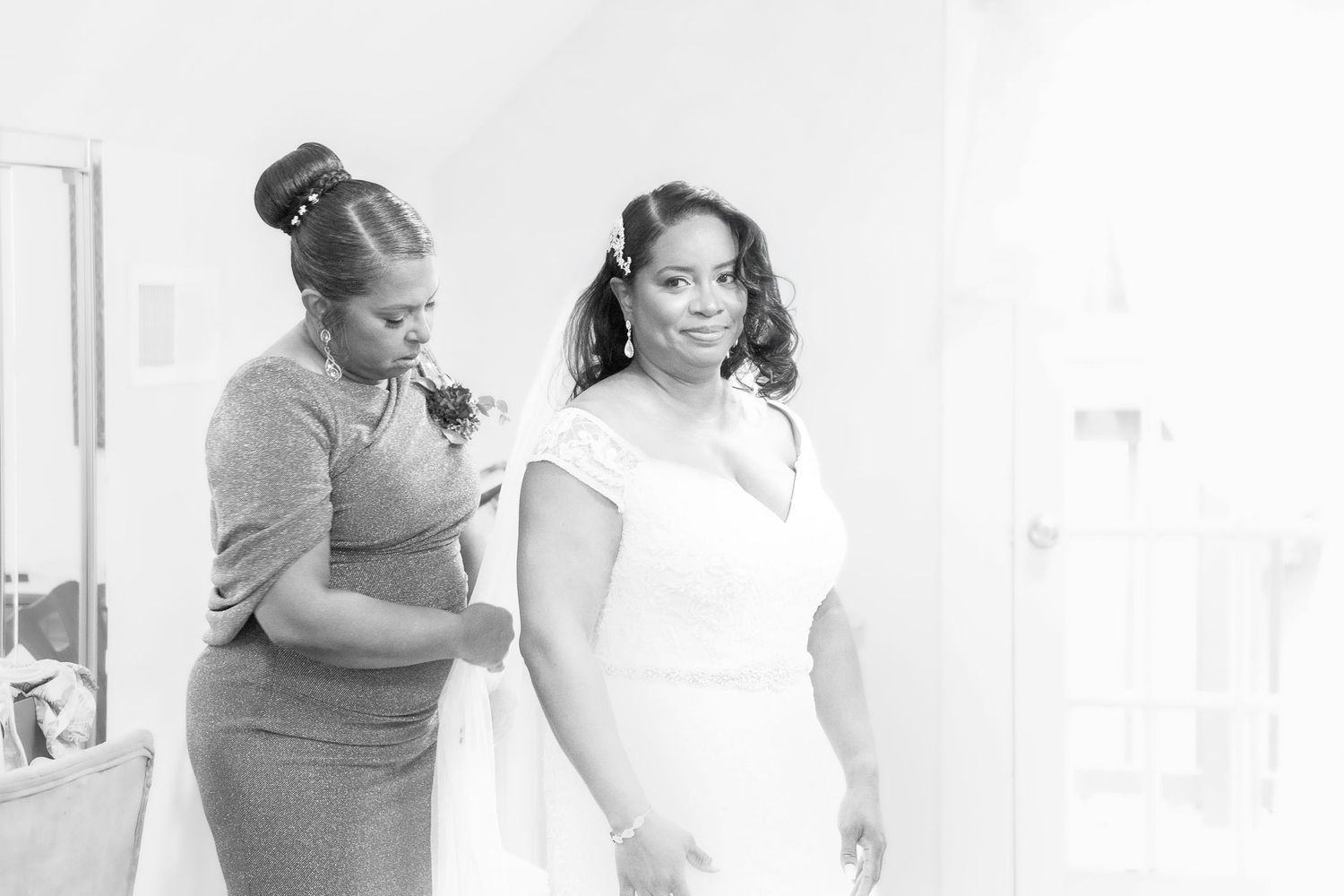A bridesmaid helps a bride in a white gown get dressed in a bright room on her wedding day.