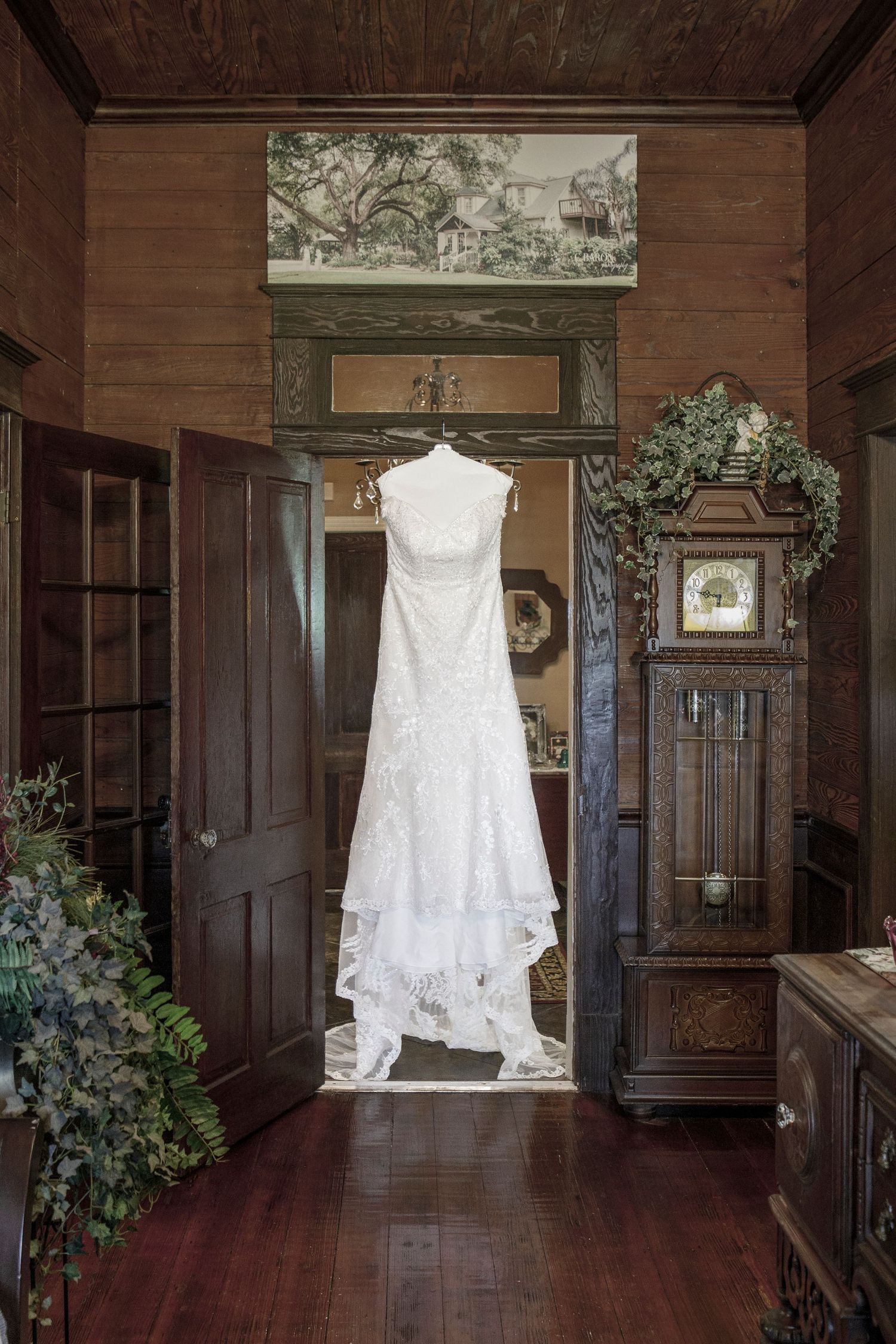 White lace wedding dress hanging in a rustic wood-paneled room with a grandfather clock and vintage decor.