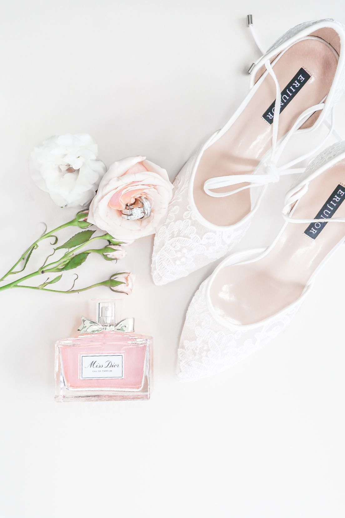 Elegant flat lay composition with pink perfume bottle, white bridal shoes, and delicate floral accents on white background.