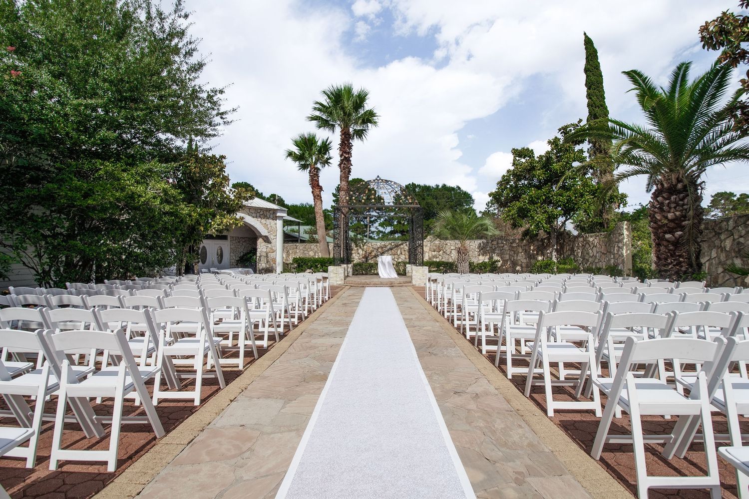 Outdoor wedding ceremony setup with white folding chairs lining a white aisle runner, flanked by palm trees and lush greenery.