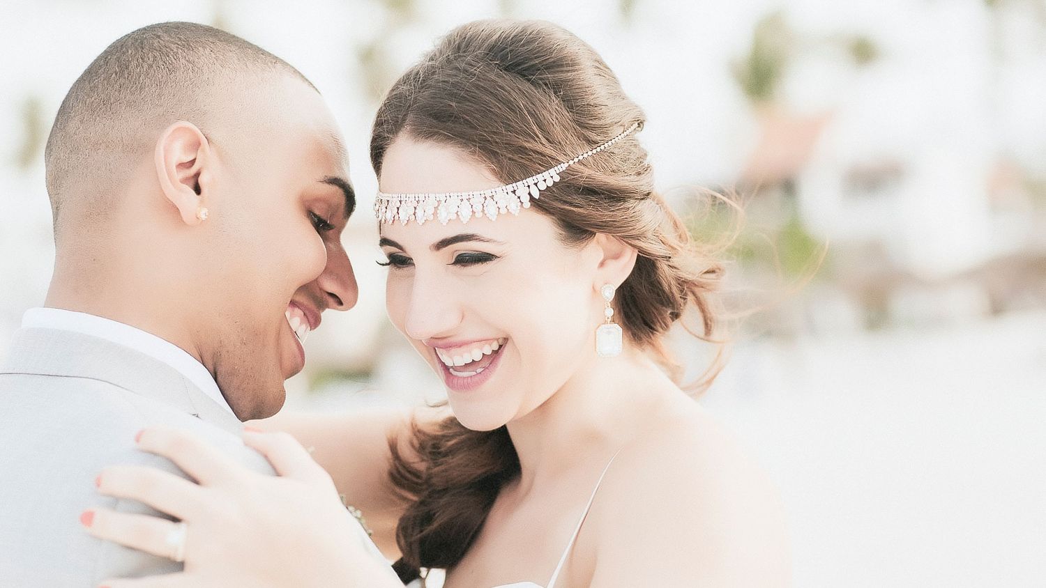 A smiling bride in a white dress and headpiece embraces her groom outdoors on their wedding day.
