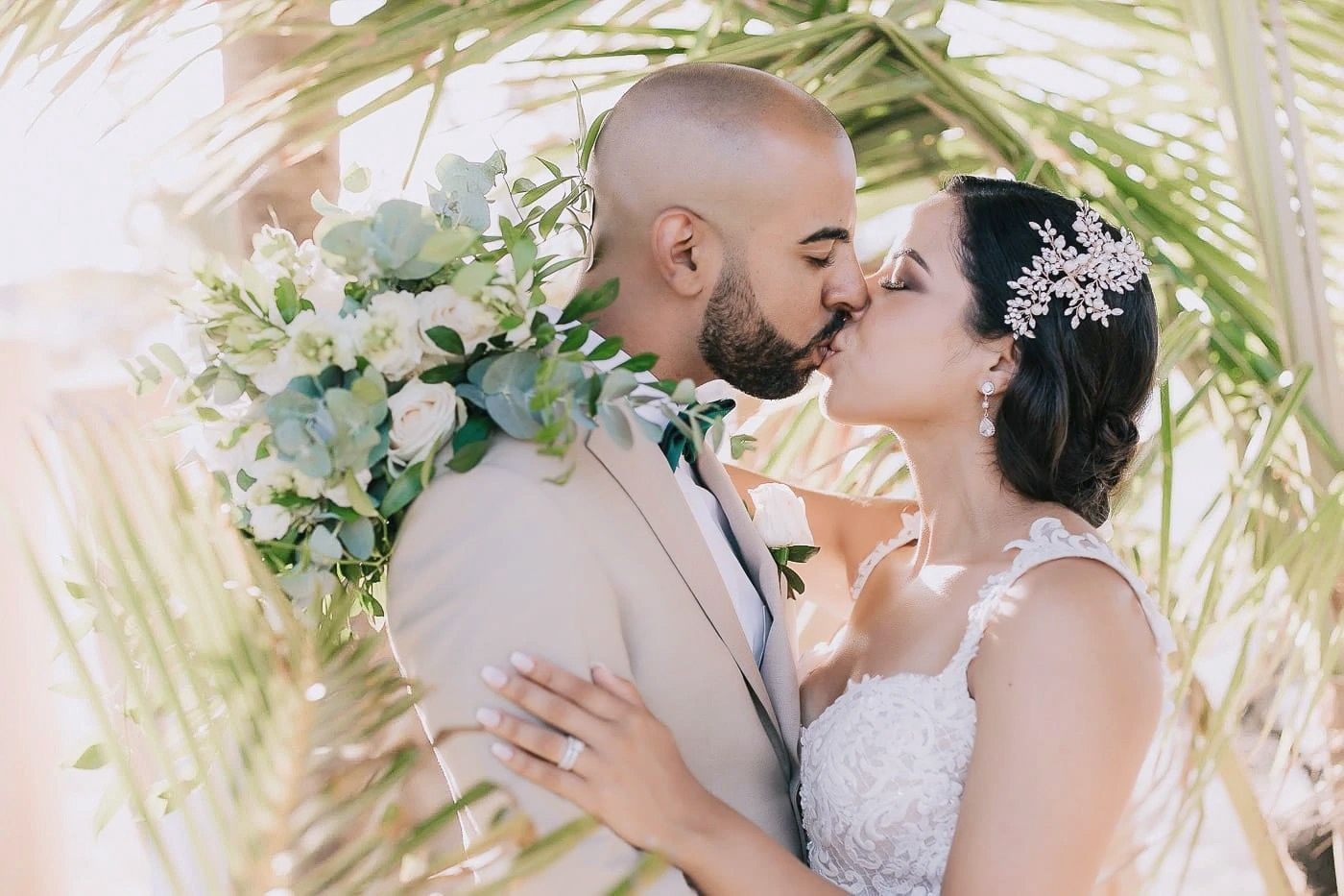 A romantic wedding couple shares a tender kiss surrounded by tropical palm fronds and white floral bouquets.
