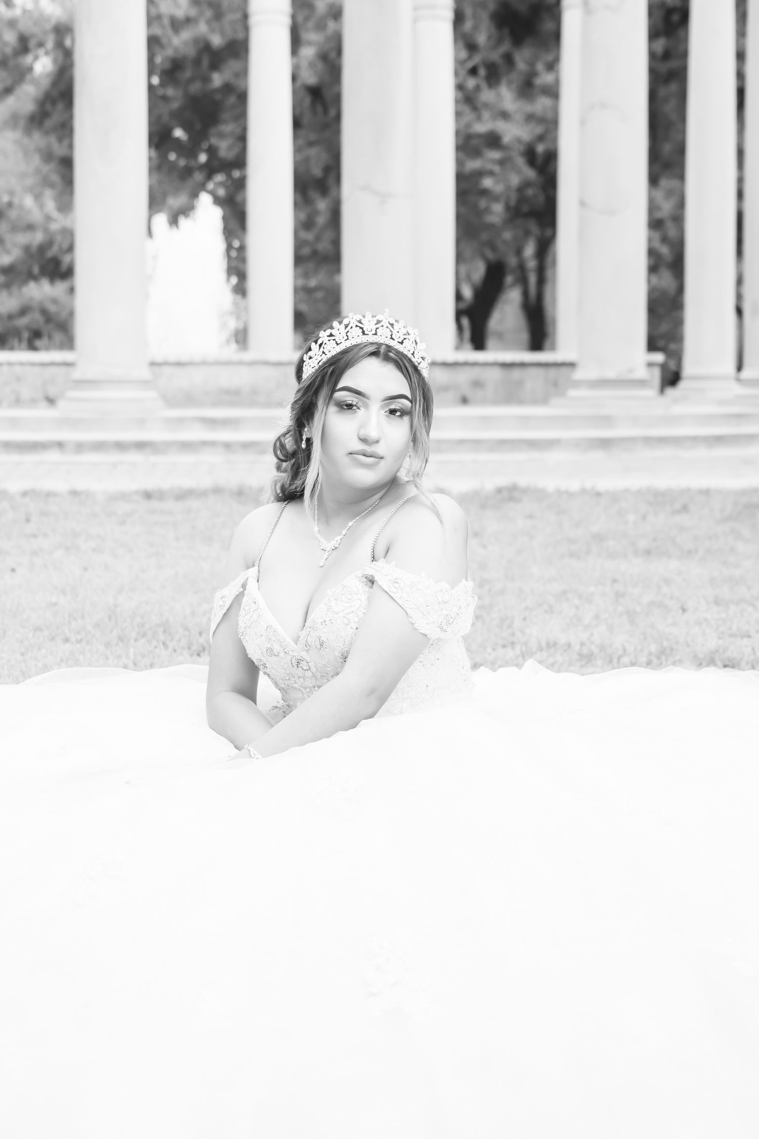 A black and white portrait of someone in an elegant white gown and tiara posing near classical columns outdoors.