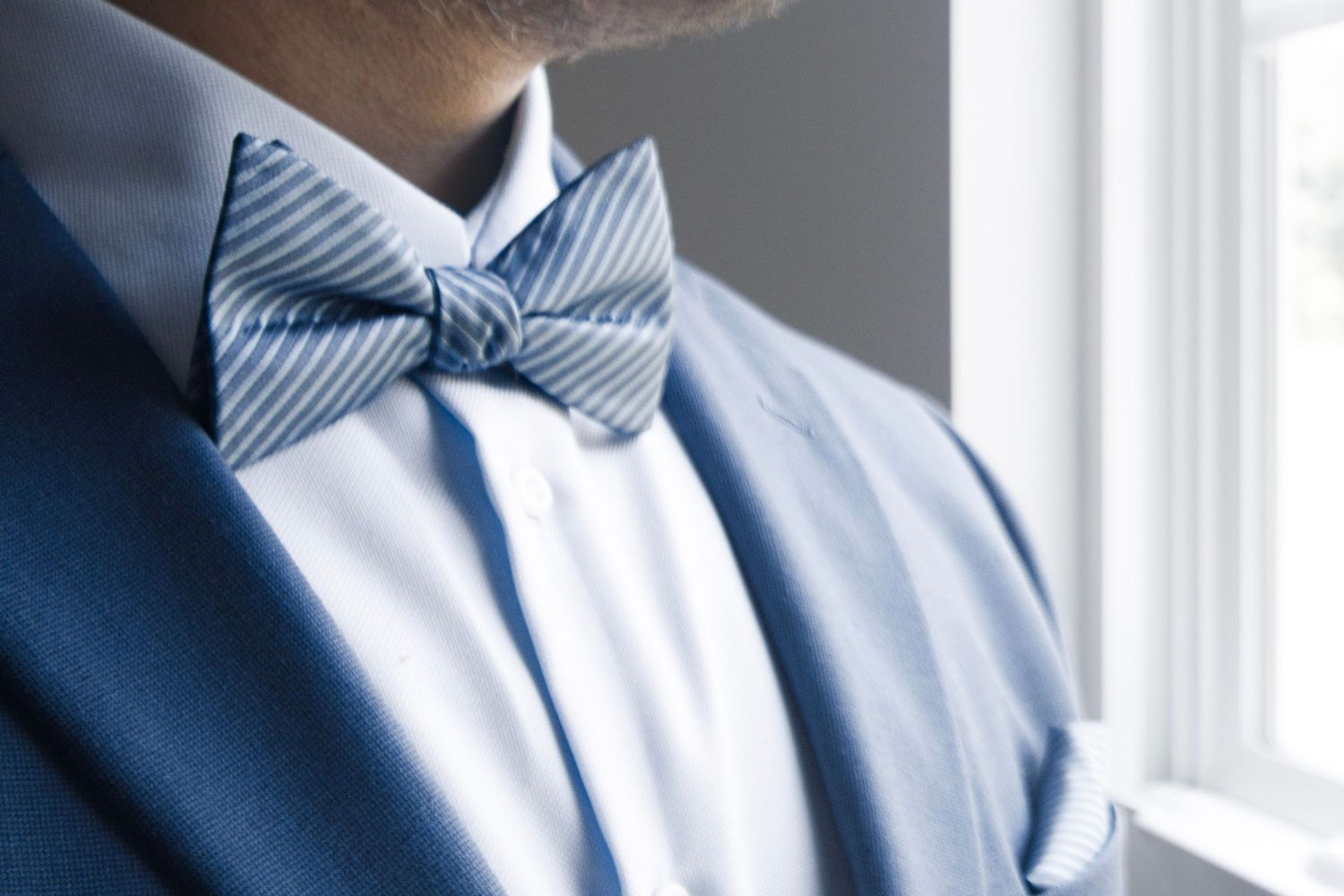 Man wearing a blue striped bow tie with a white dress shirt and blue suit jacket near a window.