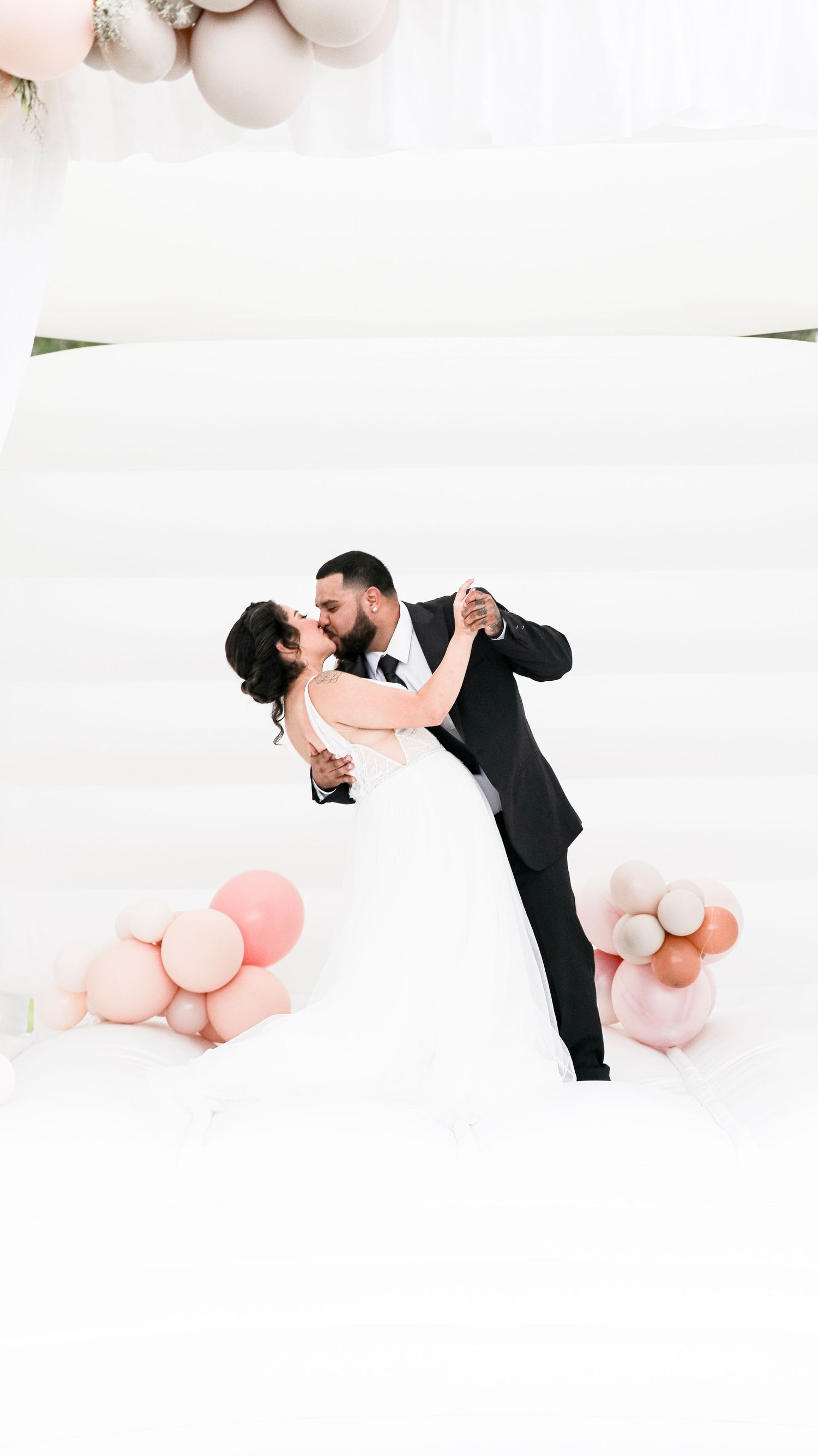 Bride and groom share a romantic dip kiss on a white dance floor surrounded by pink and neutral balloon clusters.
