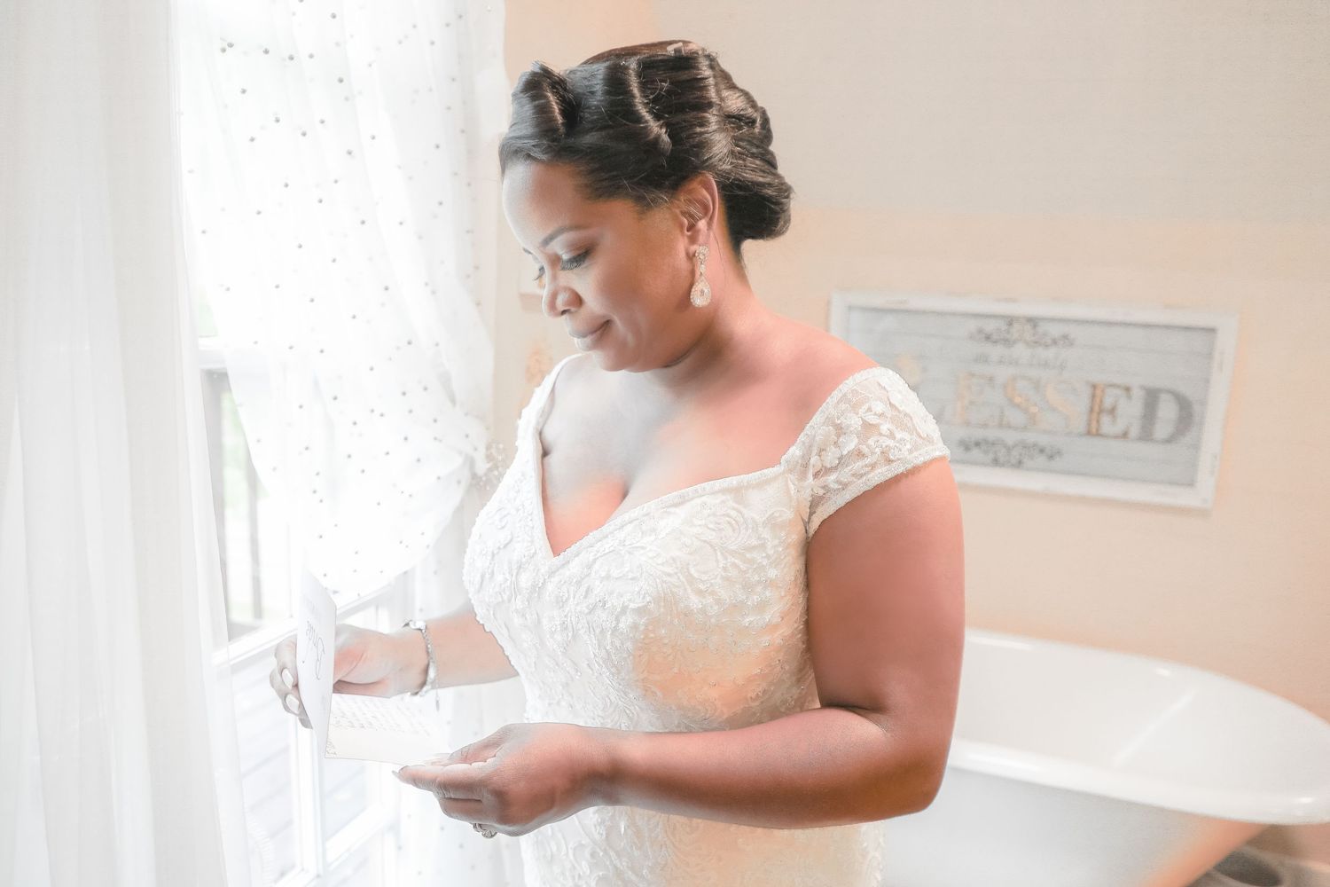 A bride in a white lace wedding dress poses near a window while getting ready for her ceremony.