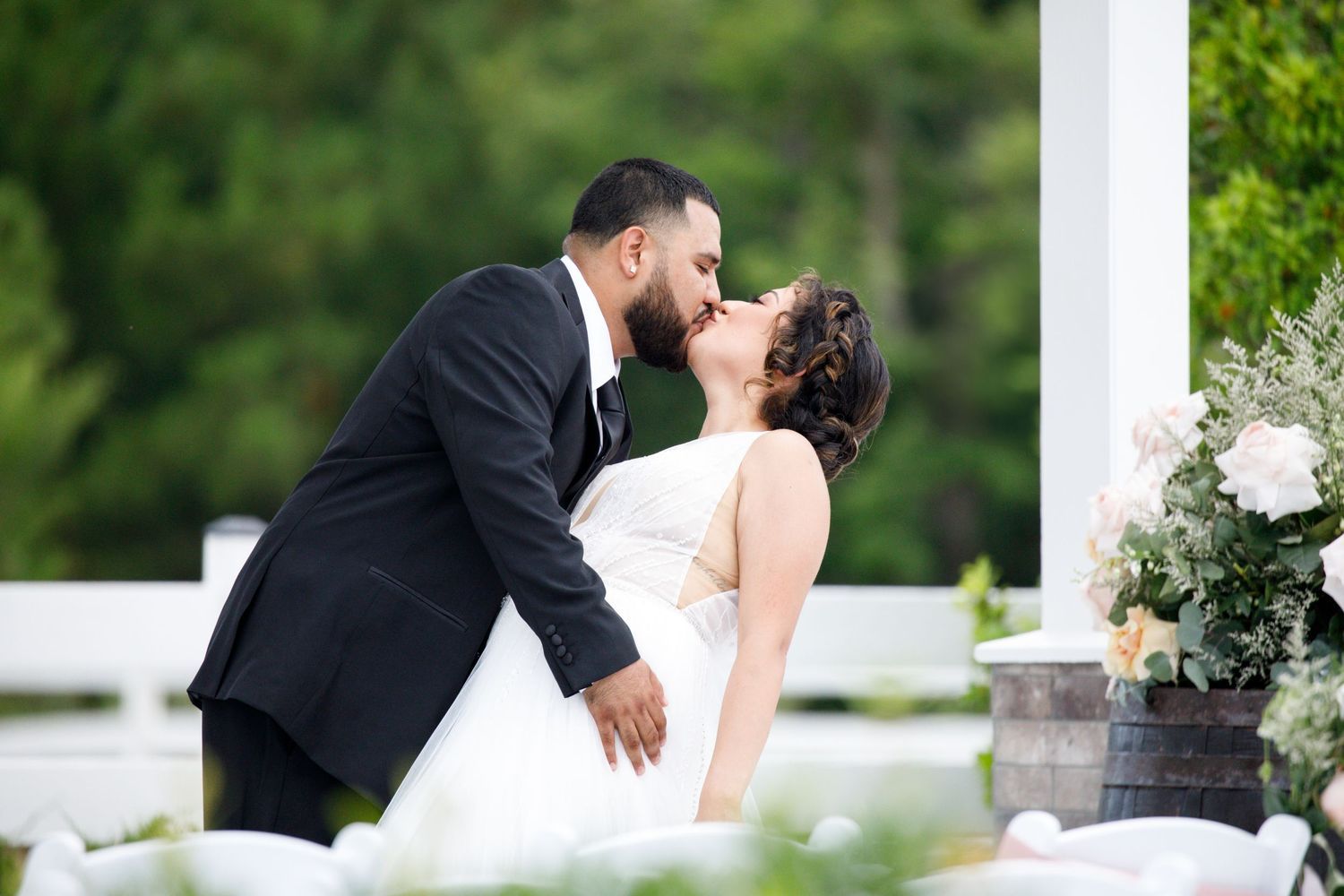 A couple shares a romantic kiss outdoors during their wedding celebration in a scenic garden setting.