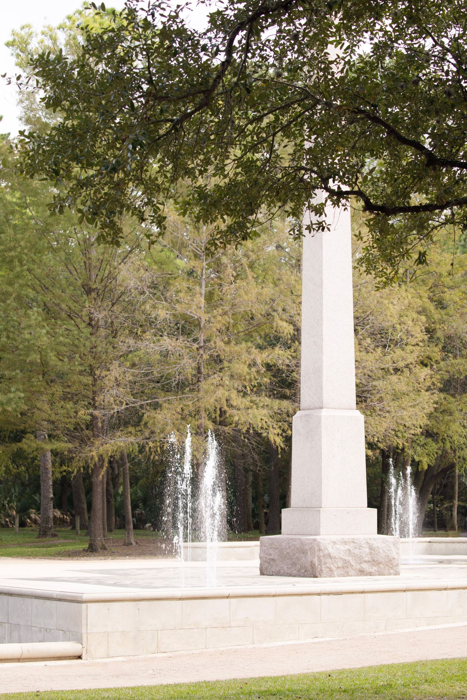 A white stone monument pillar stands in a garden plaza with active fountains and green trees in the background.