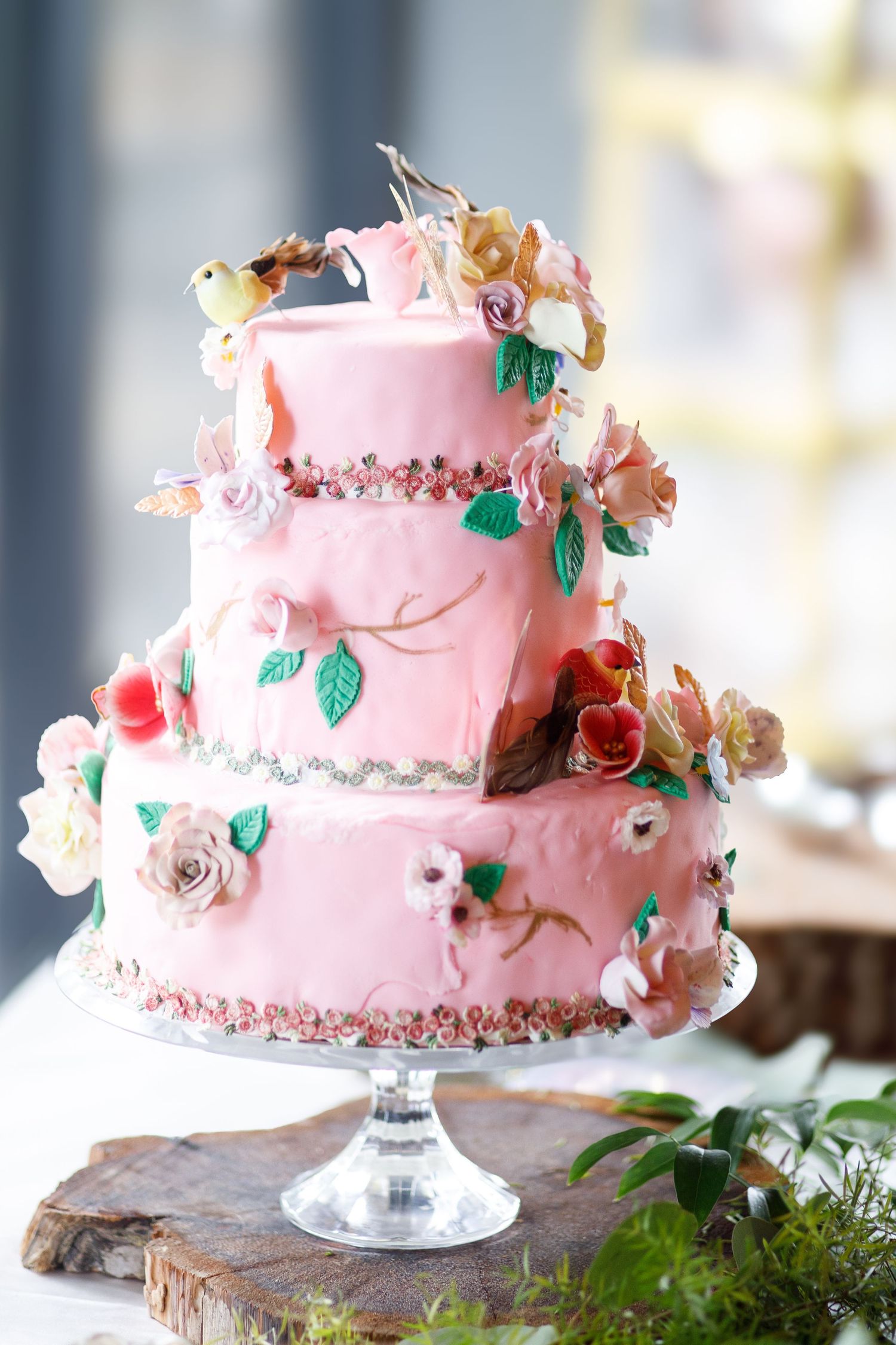 Pink wedding cake decorated with delicate roses and green leaves sits on a crystal cake stand against a soft blurred background.