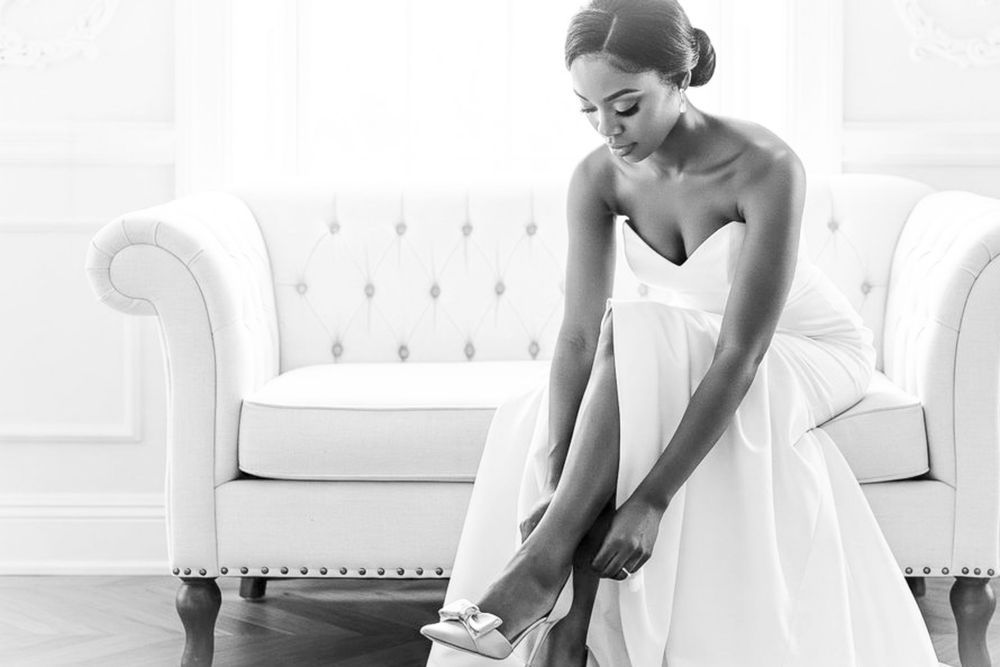 Bride in white strapless gown adjusting her wedding shoe while seated near an elegant white sofa.