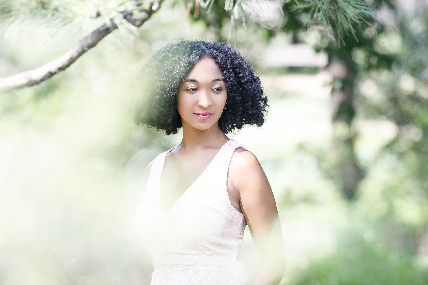 A person in a white dress poses gracefully in a dreamy outdoor setting with soft natural lighting and greenery.