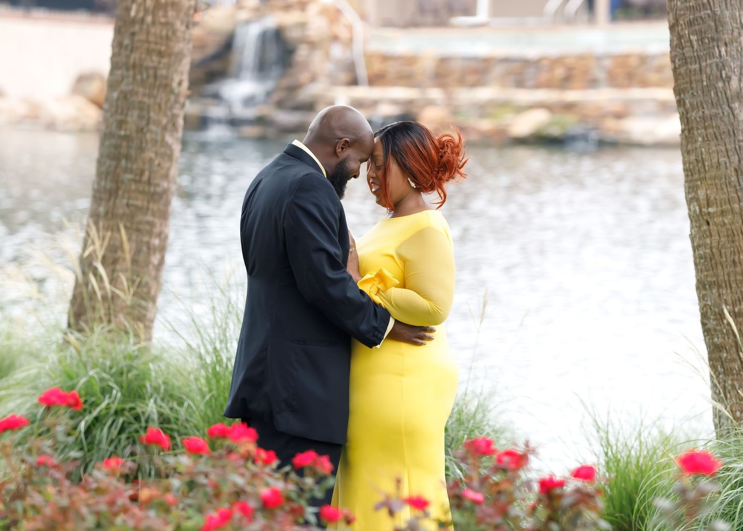 A couple shares an intimate moment in a garden with red flowers and a waterfall backdrop, with one wearing a bright yellow dress.
