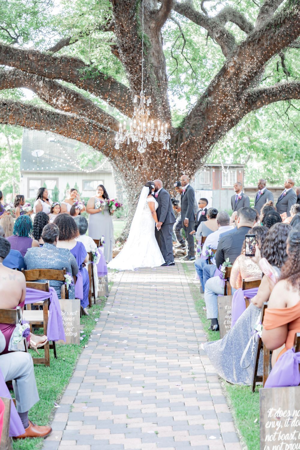Outdoor wedding ceremony under a large oak tree with purple decor and guests seated on paved pathway.