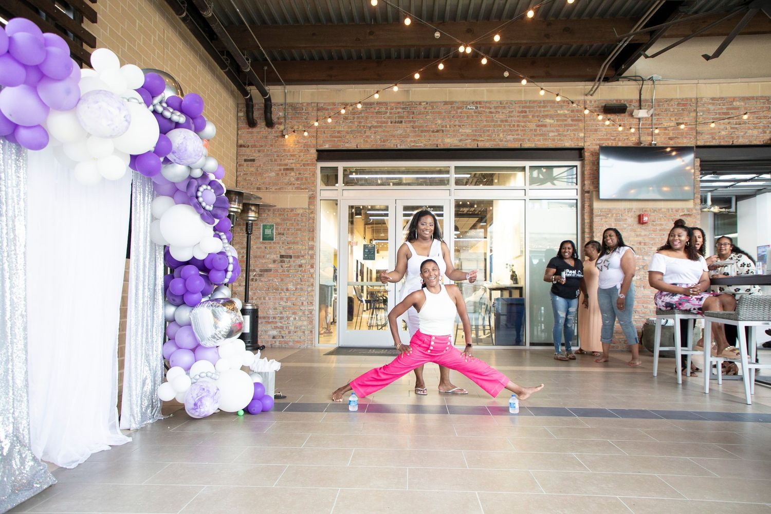 Purple and white balloons decorate an indoor event space with exposed brick walls and industrial-style ceiling.