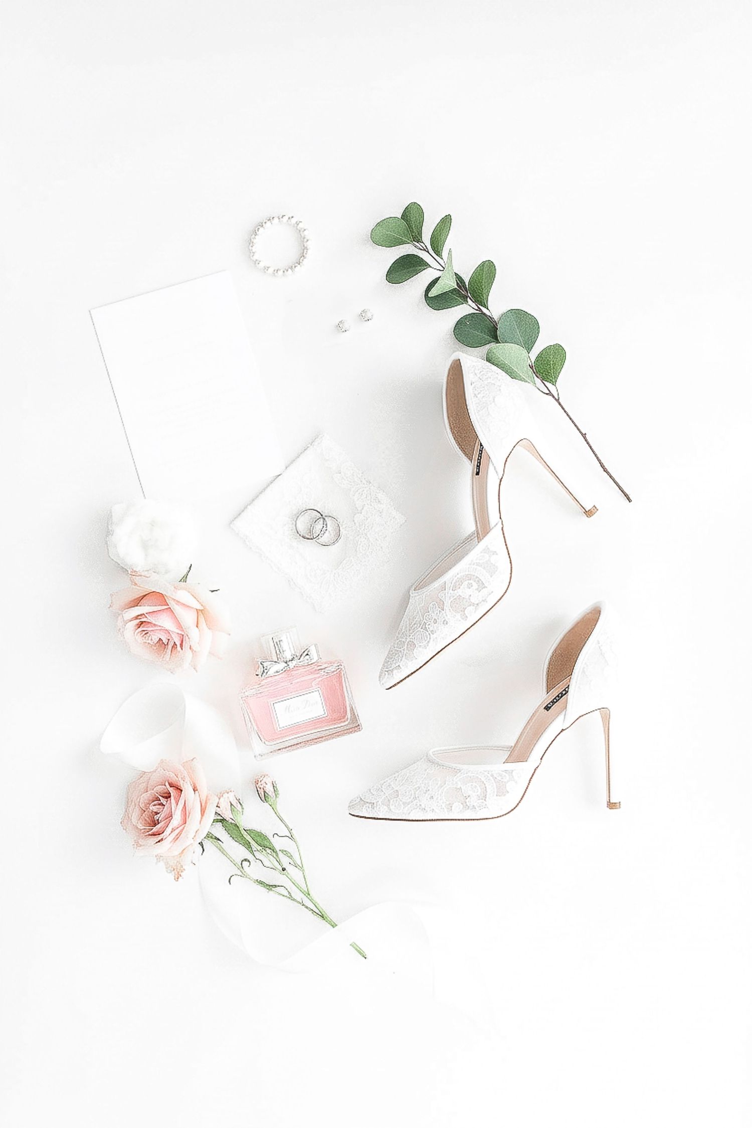 Elegant bridal flat lay with white lace heels, pink roses, perfume, wedding rings, eucalyptus, and stationery on white background.