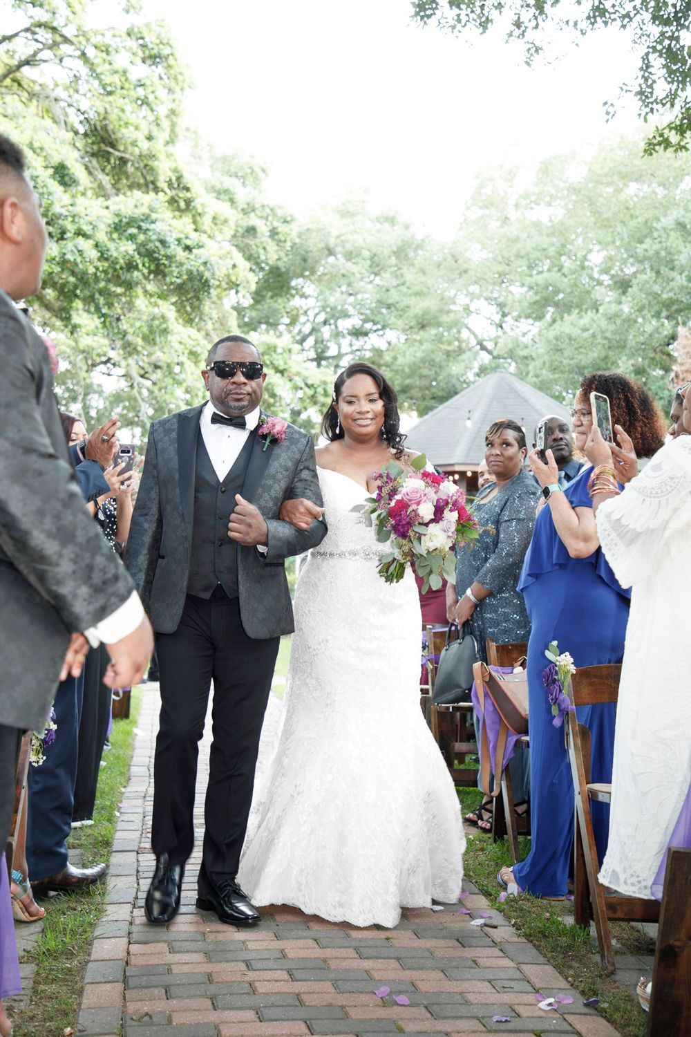 Bride and groom walk down outdoor wedding aisle, bride holding pink bouquet, guests celebrating around them.