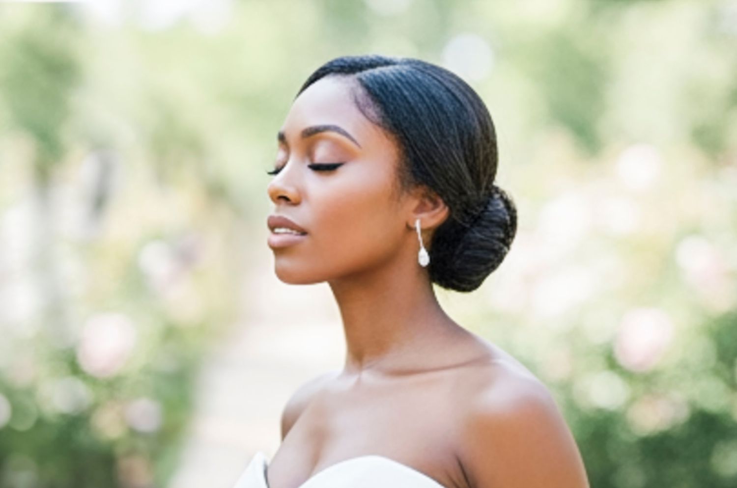 Elegant bride with updo hairstyle and drop earrings, eyes closed, wearing strapless gown outdoors.