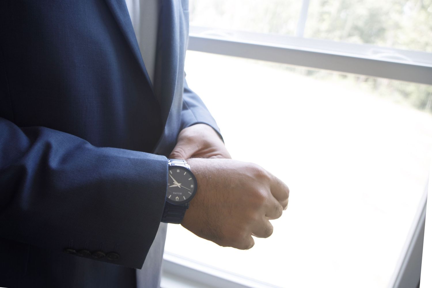 Man in navy blue suit adjusting wristwatch near window, wearing elegant black analog watch.