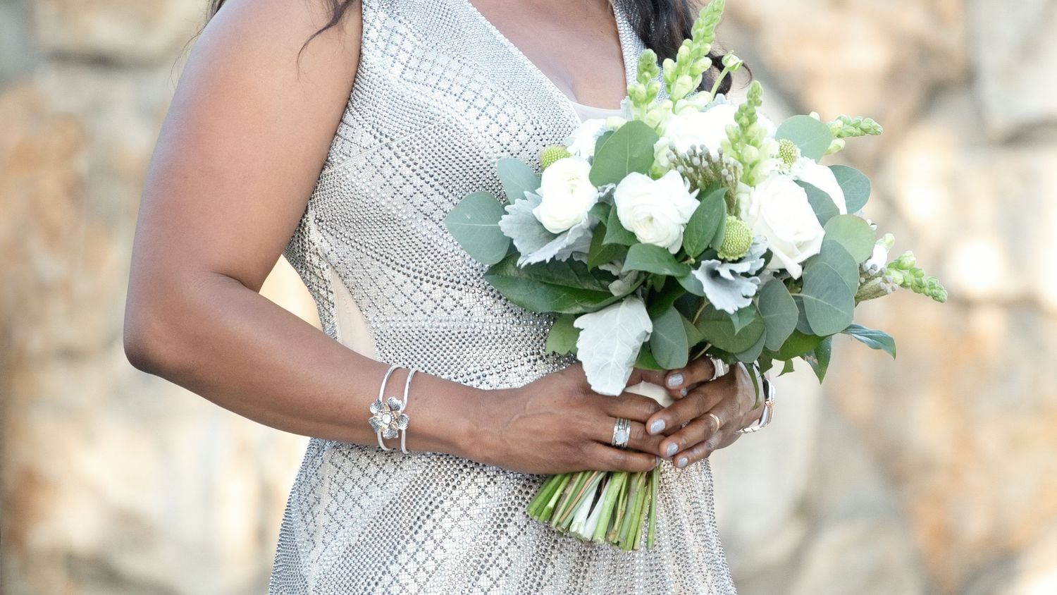 Bride in silver dress holding white rose and greenery wedding bouquet outdoors near stone wall.