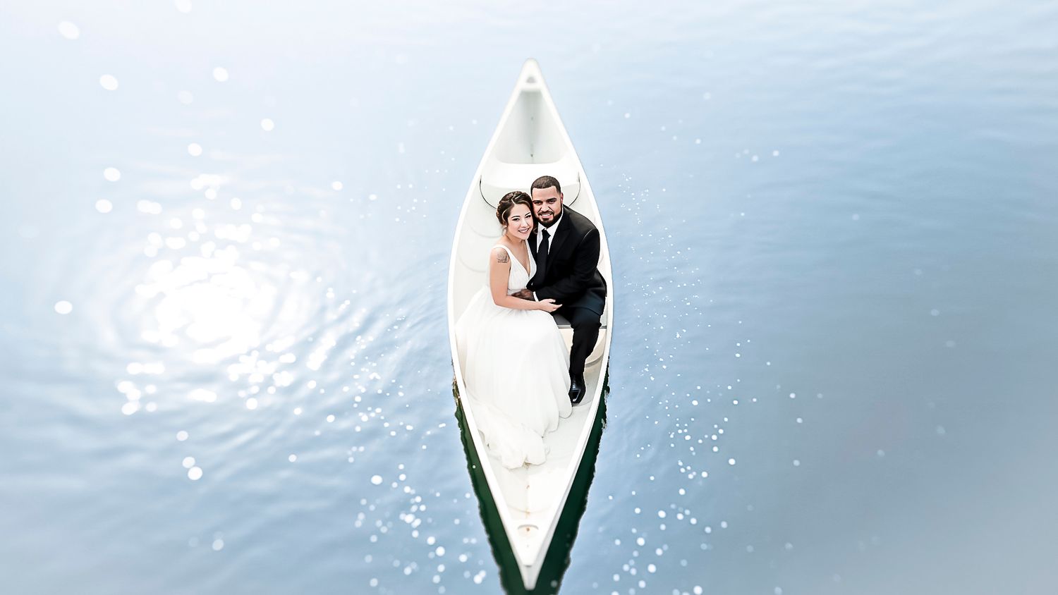 Aerial view of a bride and groom in wedding attire sitting together in a white canoe on calm blue water.