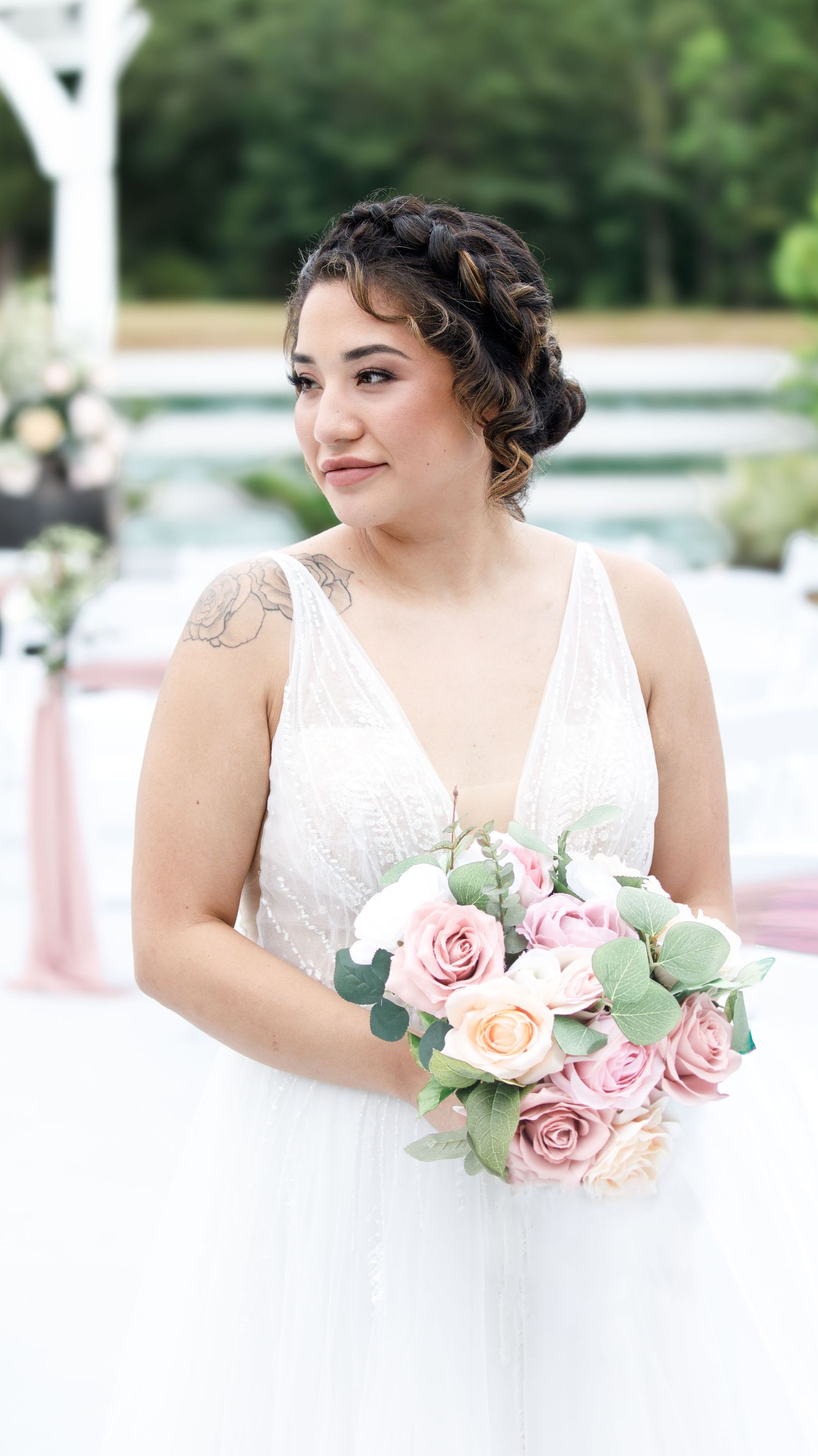 Bride in white V-neck gown holding pink rose bouquet with braided updo and shoulder tattoo at outdoor wedding.
