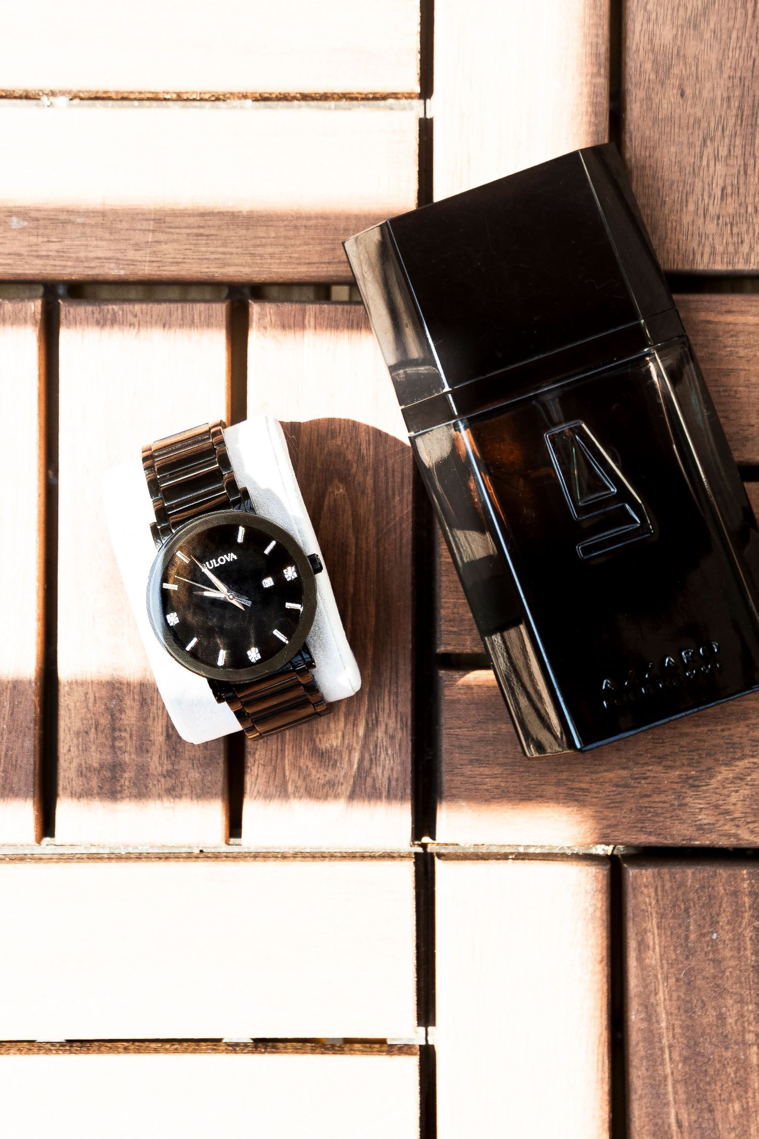 Black Bulova watch and dark cologne bottle displayed on wooden slat surface.