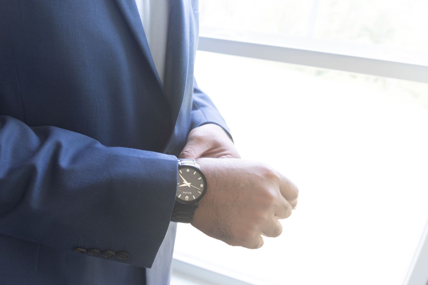 Man in blue suit adjusting his classic analog wristwatch near a bright window.