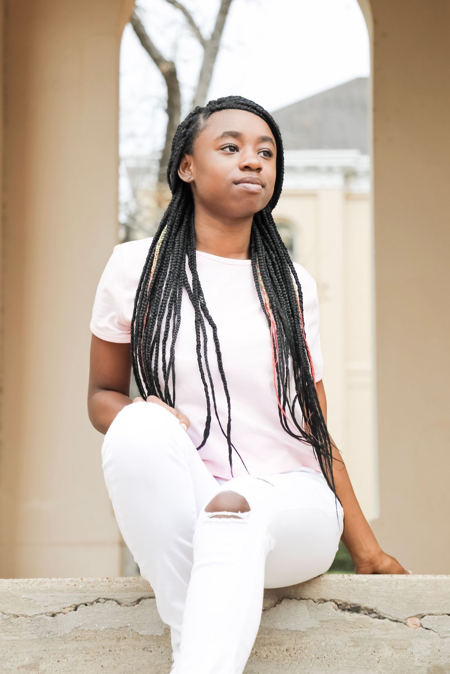 Portrait photo of a person with long braids wearing white jeans and pink t-shirt, sitting in an arched doorway.