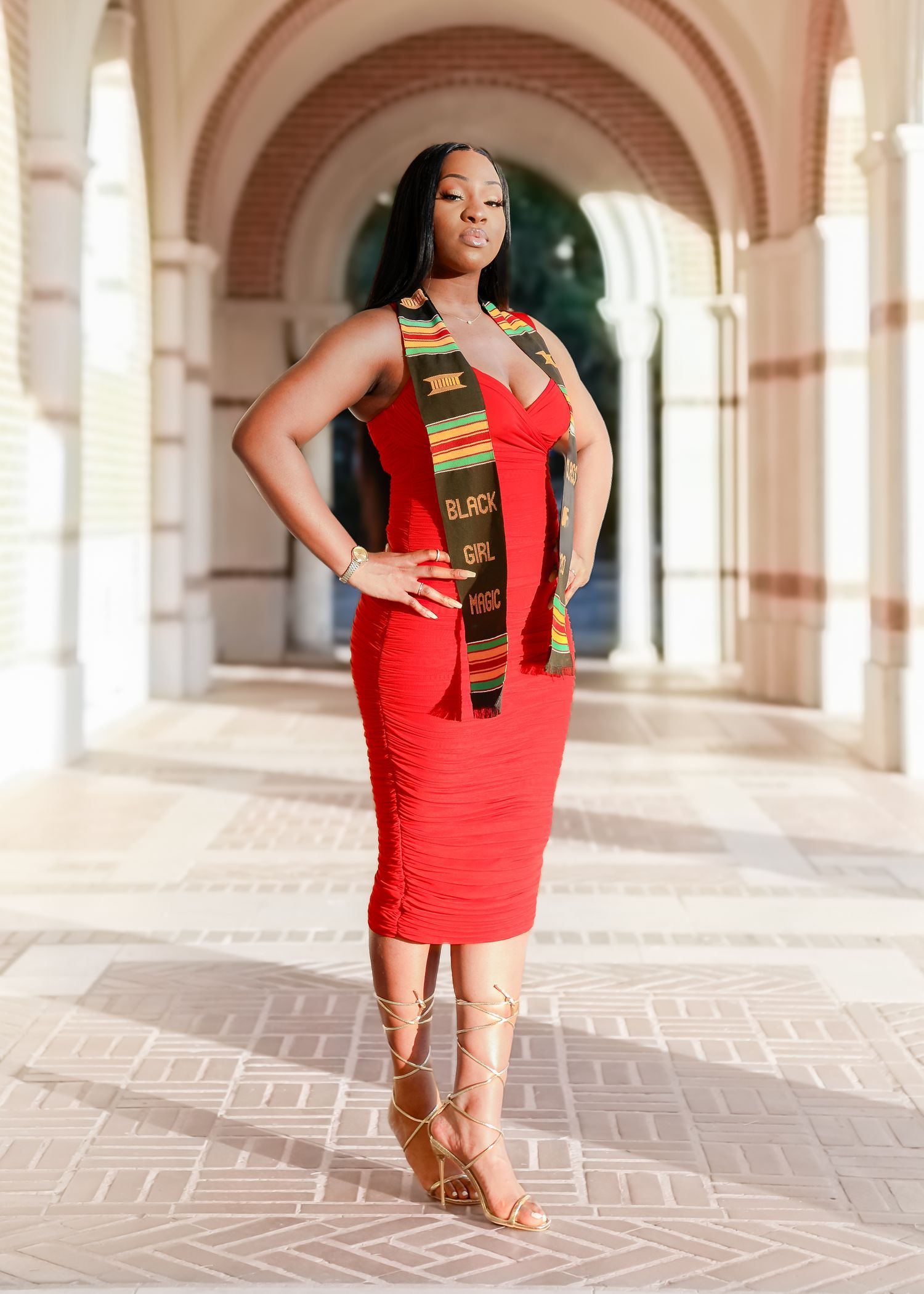 Elegant graduation photo in an arched corridor featuring a red bodycon dress and traditional kente stole graduation sash.
